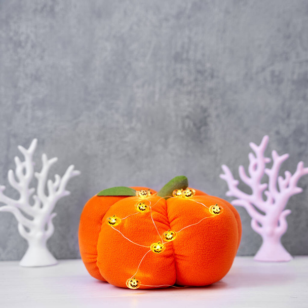 Halloween decoration toy plush pumpkin with a green tail and a garland of glowing pumpkins on a gray background - Foto, Imagem