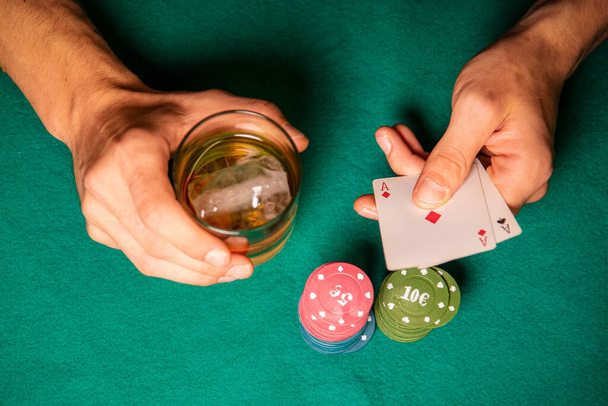 άνθρωπος με ένα ποτήρι ουίσκι στο χέρι του και μια ποσότητα χρημάτων στο τραπέζι δείχνει 2 άσους που κουβαλάει στο χέρι του - Φωτογραφία, εικόνα