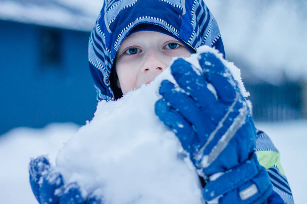 Junge in Winterjacke steht draußen und frisst Schnee mit einem großen Klumpen Schnee in der Hand - Foto, Bild