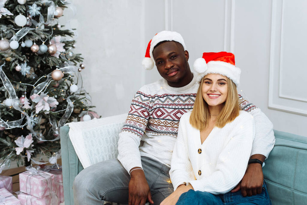 A man and a woman are looking at the camera, smiling in santa hats and sitting on a sofa in a bright interior next to a Christmas tree - Foto, Bild