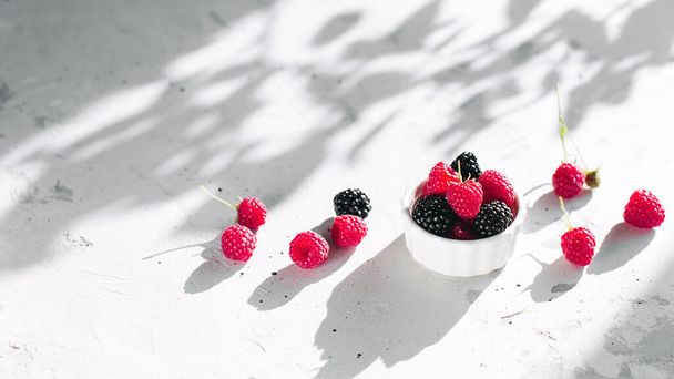 White bowl full with raspberries, blackberry on gray concrete table with leaves, tree branches shadow. Healthy eating concept. Eco, bio farming. Fresh tasty berries on grey background. Quality photo - Zdjęcie, obraz