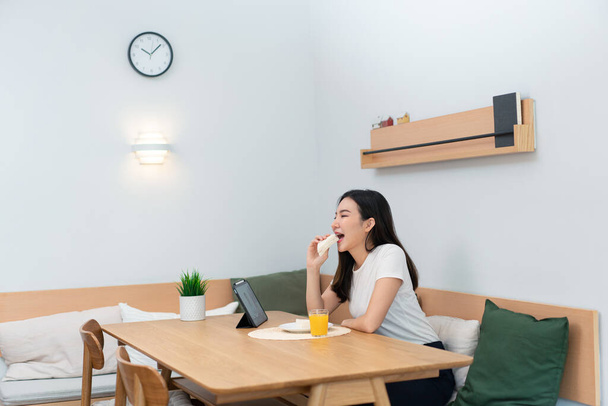 Olohuoneen käsite naispuolinen aikuinen nauttii syöminen voileipiä ja appelsiinimehua katsella online-median työ tauko. - Valokuva, kuva
