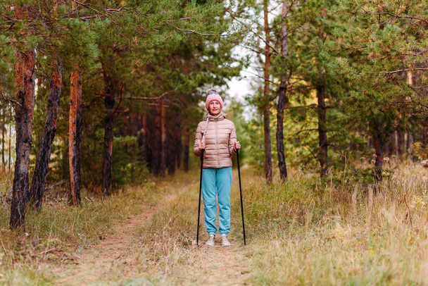 an elderly sixty-year-old woman in sportswear is engaged in Nordic walking with sticks in the forest in the autumn - Φωτογραφία, εικόνα