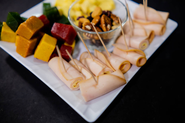 Рулети з шинки, нарізана фруктова паста, кубики сиру та горіхи на квадратній білій тарілці
 - Фото, зображення
