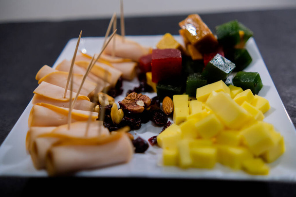 Plate of ham rolls, fruit paste, cheese cubes, and nuts on black surface - Foto, imagen