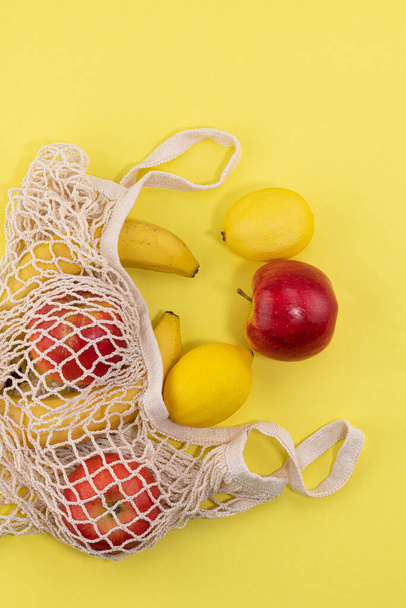 mesh shopping bag with organic fruit on yellow background. Cotton shopping bag with Lemons, bananas, apples. Caring for the environment. Zero waste concept. No plastic. Top view with copy space. - Foto, imagen