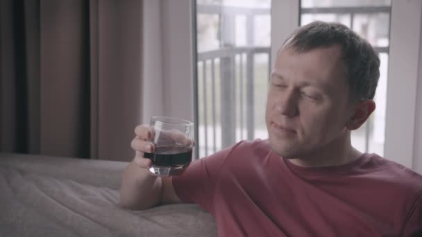 Грустный человек сидит на диване со стаканом напитка в руке, на заднем плане окна - Кадры, видео