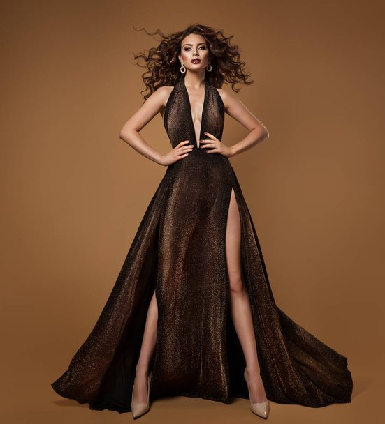 High Fashion Model in Black Dress with Golden Glitters over Beige. Luxury Brunette Woman in Sexy Evening Gown with Curly Hair fluttering on Wind - Fotó, kép