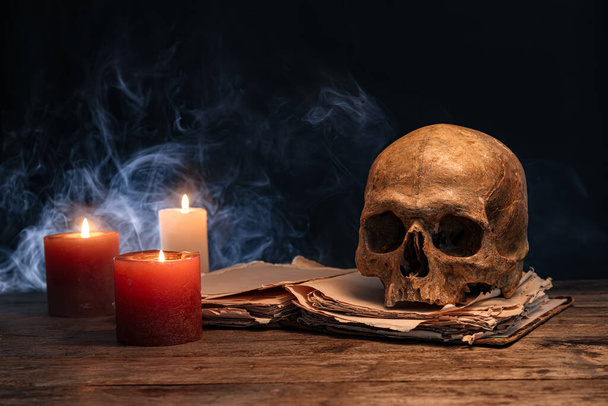 Human skull with book and glowing candles on table against dark background - 写真・画像
