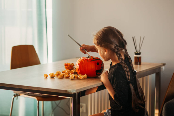 A little girl decorates a pumpkin in a room for Halloween. Kids decorate home. Children carve pumpkin. Spooky Halloween fun - Photo, Image