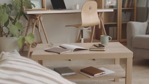Tilting-up no people shot przytulny minimalistyczny salon z drewnianym stołem kawowym, krzesło i biblioteczka ze szklanymi drzwiami, rośliny domowe i laptop na biurku - Materiał filmowy, wideo