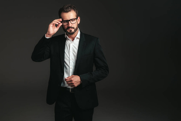 Portrait of handsome confident stylish hipster lambersexual model. Sexy modern man dressed in elegant black suit. Fashion male posing in studio on dark background. In spectacles - Φωτογραφία, εικόνα