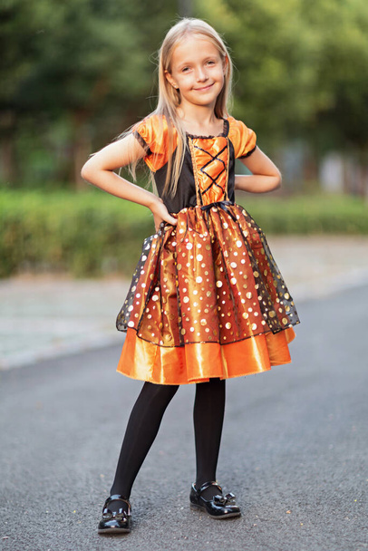 Lifestyle portrait of Happy Little caucasian Girl with blonde hair seven years old in black orange costume of which celebrating Halloween alone outdoor during Coronavirus covid-19 pandemic quarantine. - Fotó, kép