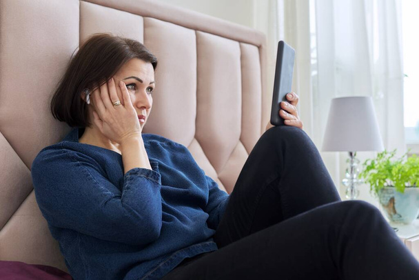 Smutna kobieta w średnim wieku w słuchawkach patrząc w ekran smartfona. - Zdjęcie, obraz