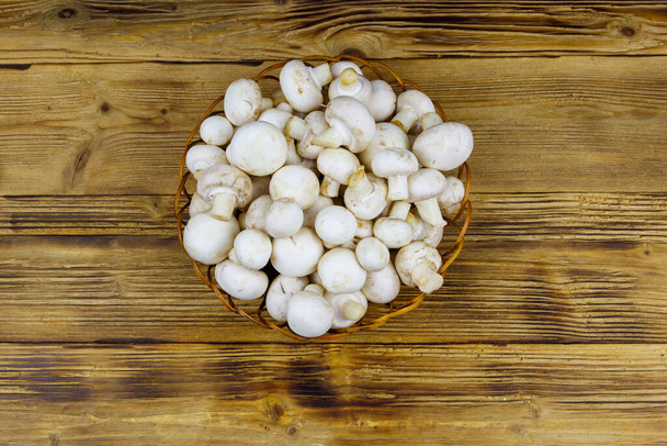 Fresh champignon mushrooms in wicker basket on the wooden table. Top view - Fotografie, Obrázek