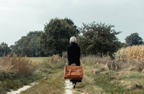 Stylowa kobieta w czarnym płaszczu i walizce spacerując po wiejskiej drodze - Zdjęcie, obraz