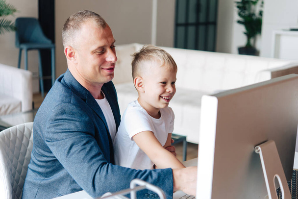 Padre feliz se sienta y trabaja en la computadora, a su lado hay un niño preescolar feliz, mirando al monitor y sonriendo. - Foto, Imagen