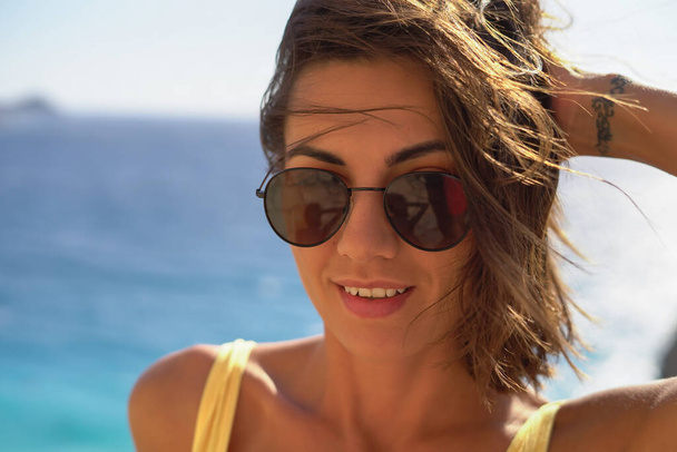 Νεαρή γυναίκα με γυαλιά ηλίου σε μια υπέροχη διάθεση στις διακοπές με φόντο τη θάλασσα και την παραλία απολαμβάνοντας μια ζεστή καλοκαιρινή μέρα - Φωτογραφία, εικόνα