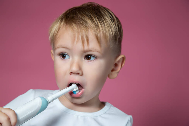 Smiling caucasian little boy cleaning his teeth with electric sonic toothbrush on pink background. - Valokuva, kuva