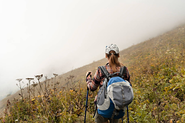 再生されたシャツの若い女性のリアビュー大きなバックパックとトレッキングポール霧の中で山道ハイキングに沿って歩くと,健康的なアクティブライフスタイル,デジタルデトックス,週末の活動 - 写真・画像