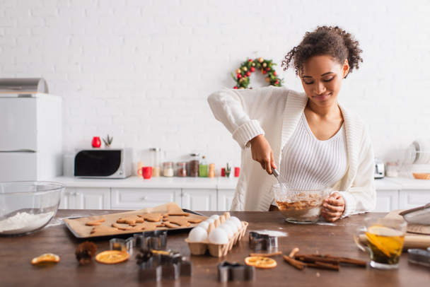 Mujer afroamericana sonriente cocinando cerca de ingredientes y galletas de Navidad en la cocina  - Foto, Imagen