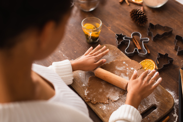 Vue grand angle de la femme afro-américaine floue rouler la pâte près des emporte-pièces de Noël et le thé dans la cuisine  - Photo, image