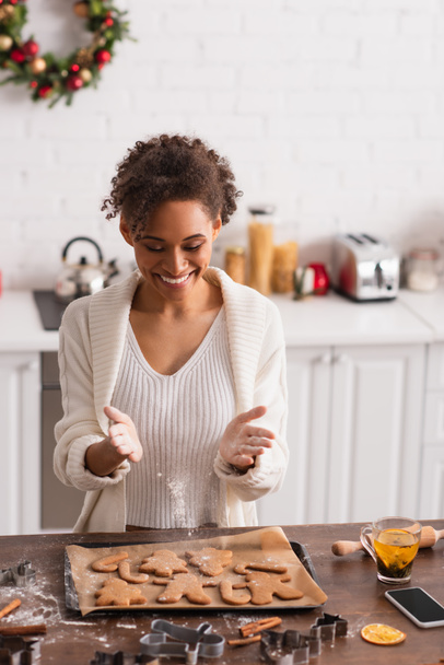 笑顔アフリカ系アメリカ人女性がスマートフォンやお茶の近くのクリスマスクッキーに小麦粉を注ぐ  - 写真・画像