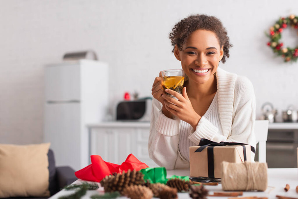 Donna afro-americana sorridente che tiene il tè vicino al presente e all'arredamento a casa  - Foto, immagini