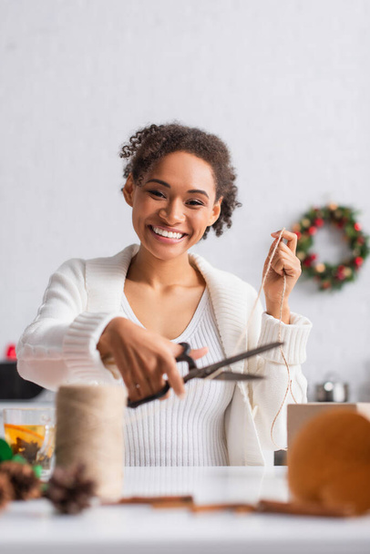Positive african american woman cutting twine near gift and cup in kitchen  - Photo, Image
