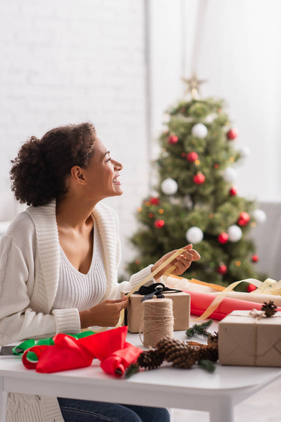 Vista lateral de la alegre mujer afroamericana decorando regalo cerca del borroso árbol de navidad  - Foto, imagen
