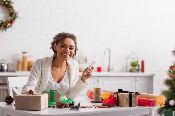 Позитивна афро-американська жінка, яка тримає мобільний біля подарунків, кристама декор і чай.  - Фото, зображення
