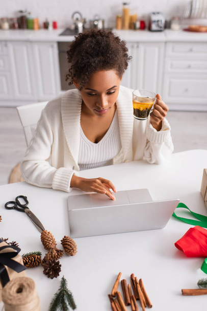 アフリカ系アメリカ人女性保持お茶と使用ノートパソコン近くクリスマス装飾で自宅  - 写真・画像