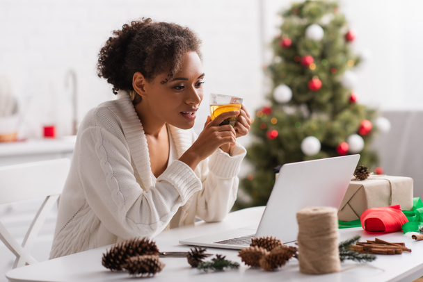 アフリカ系アメリカ人女性ノートパソコンの近くでお茶を保持し、現在とクリスマスの装飾  - 写真・画像