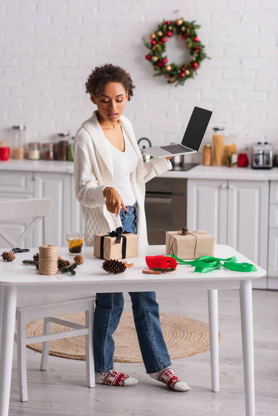 Femme afro-américaine avec ordinateur portable pointant vers cadeau près de décor de Noël à la maison  - Photo, image