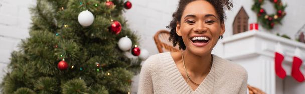 Cheerful african american woman looking at camera near blurred christmas tree at home, banner  - 写真・画像