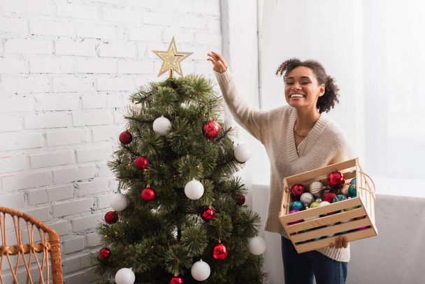 Alegre mujer afroamericana sosteniendo caja con bolas de Navidad cerca del árbol en casa  - Foto, Imagen