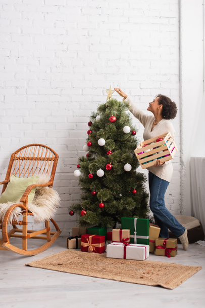 Oldalnézet pozitív afro-amerikai nő díszítő karácsonyfa közelében ajándékok otthon  - Fotó, kép