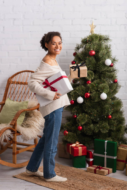Smiling african american woman looking at camera while holding presents near christmas tree at home  - Foto, Imagen