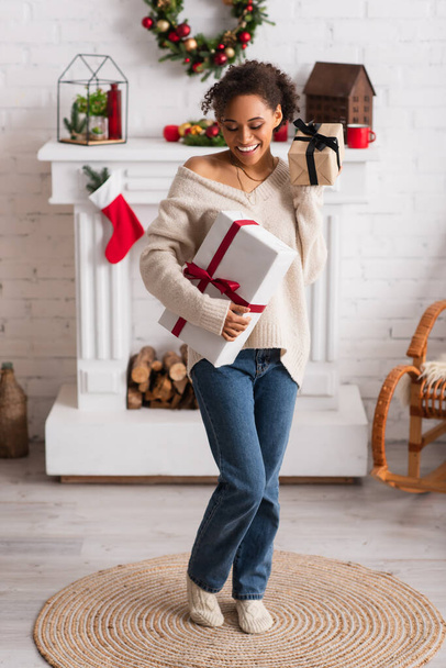 Cheerful african american woman holding presents near fireplace and christmas wreath  - Photo, Image
