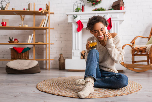 Smiling african american woman with cup of tea talking on cellphone near decorated fireplace at home  - Photo, Image