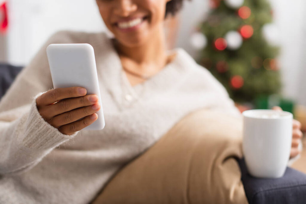 Gewassen weergave van smartphone in de hand van wazig Afrikaans-Amerikaanse vrouw met kopje thuis  - Foto, afbeelding
