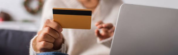 Cropped view of credit card in hand of blurred african american woman near laptop at home, banner  - 写真・画像
