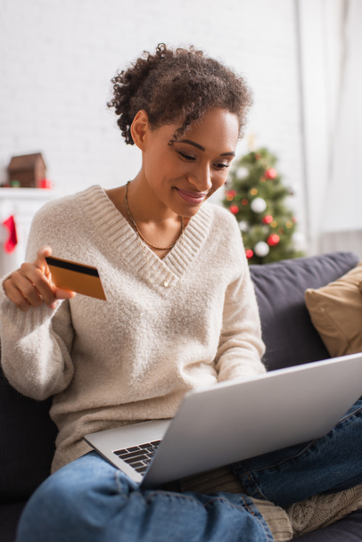 Sorrindo afro-americana segurando cartão de crédito e usando laptop durante o Natal em casa  - Foto, Imagem