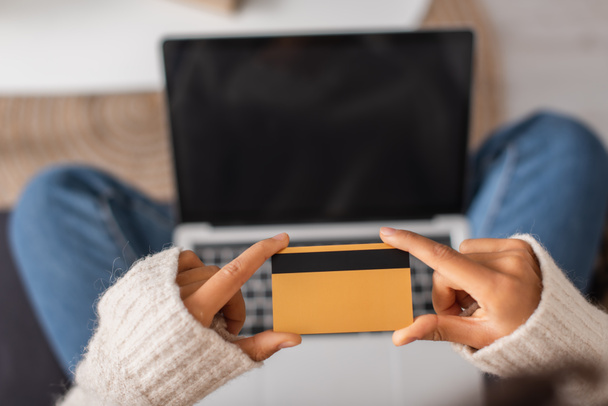 Vue recadrée d'une femme afro-américaine tenant une carte de crédit près d'un ordinateur portable flou à la maison - Photo, image