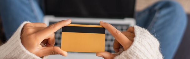 Vue recadrée d'une femme afro-américaine tenant une carte de crédit près d'un ordinateur portable flou, bannière  - Photo, image