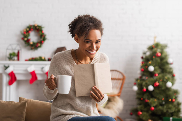 Femme afro-américaine souriante avec tasse et livre regardant la caméra pendant Noël à la maison  - Photo, image