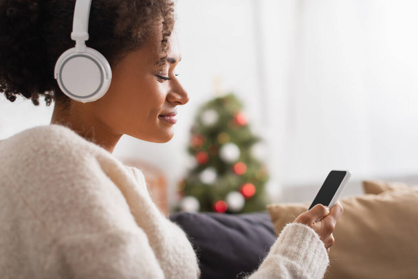 Side view of african american woman in headphones using cellphone during christmas at home  - Photo, Image