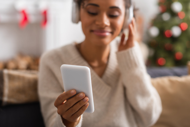 Homályos afro-amerikai nő fülhallgatóban okostelefont használ karácsonykor otthon  - Fotó, kép