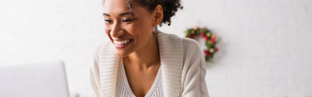 Mulher afro-americana positiva usando laptop durante o Natal em casa, banner  - Foto, Imagem