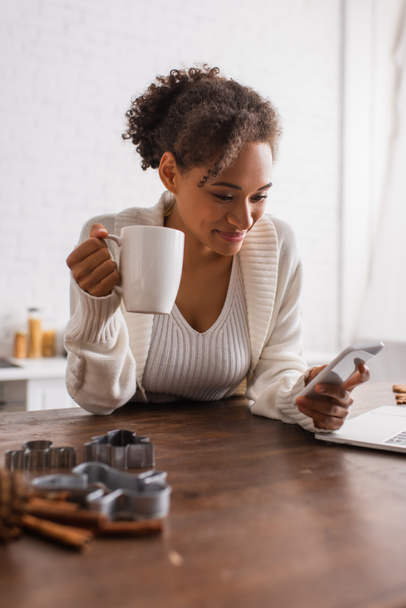 African american woman with cup using smartphone near laptop and cookie cutters in kitchen  - Photo, Image